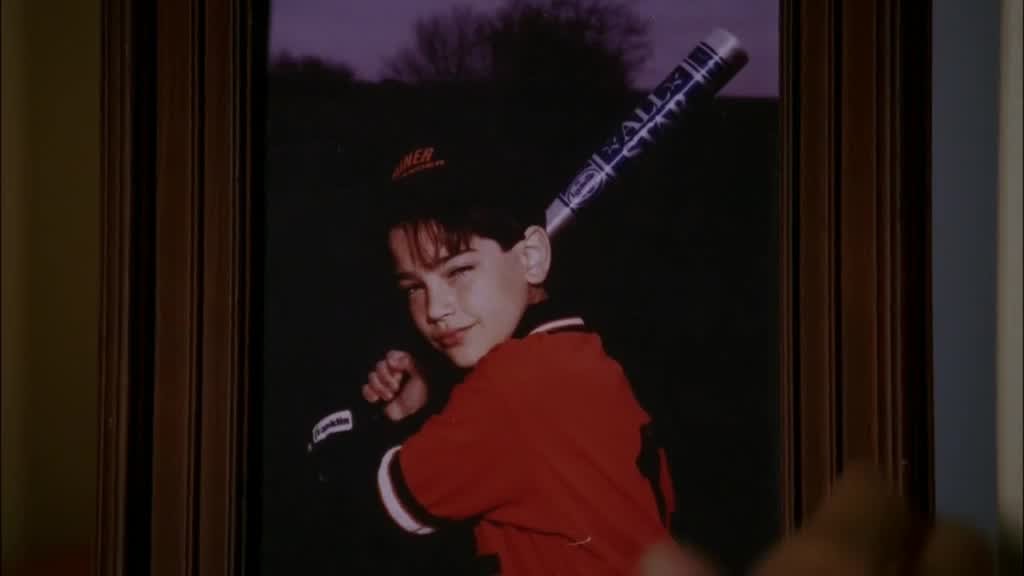 GABRIELLE catches sight of a picture of a younger JOHN playing baseball next to .jpg